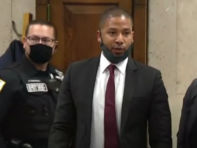 Jussie Smollett is on a suit with a black mask on and there is a cop behind him.